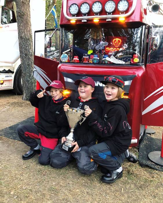 Tre barn framför en liten röd lastbil med en guldig pokal i handen.
